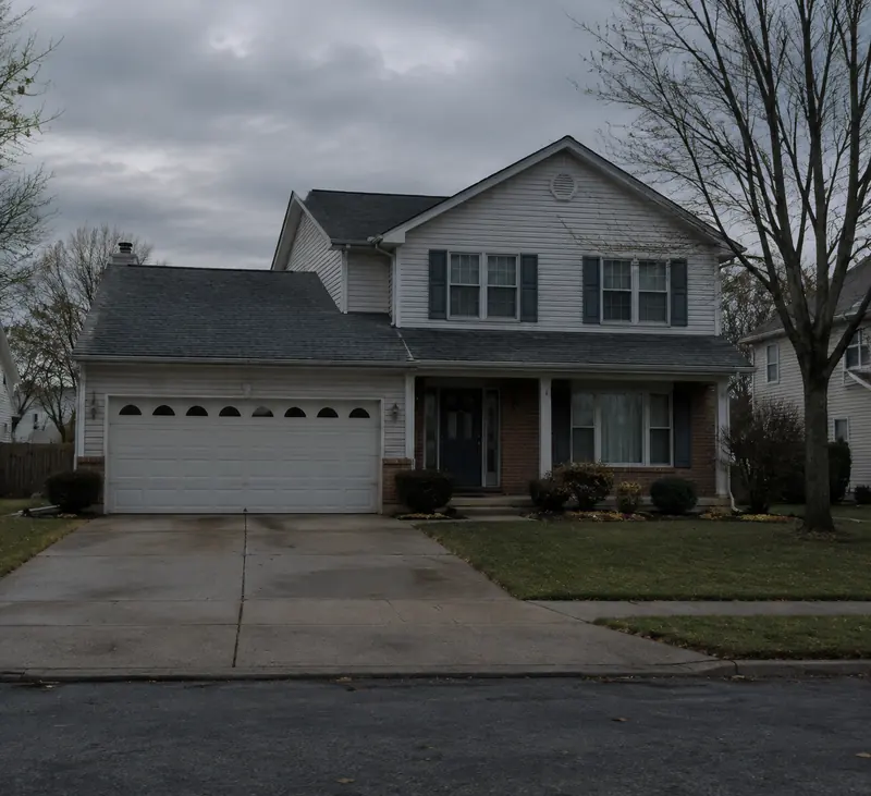 Une maison de banlieue à étage, bardage clair et volets sombres, garage double et pelouse tondue, sous un ciel gris d'hiver. Les arbres sont nus.